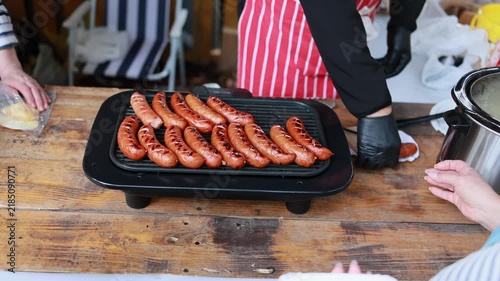 Man in black gloves grilling sausages on electric grill at outdoor street food market. Vendor in striped apron cooking grilled sausages and serving food to customers on wooden table