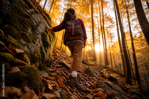 Woman hiking uphill on a rocky trail in an autumn forest. Female hiker with backpack exploring sunny woods with fall foliage. Active outdoor lifestyle and adventure concept