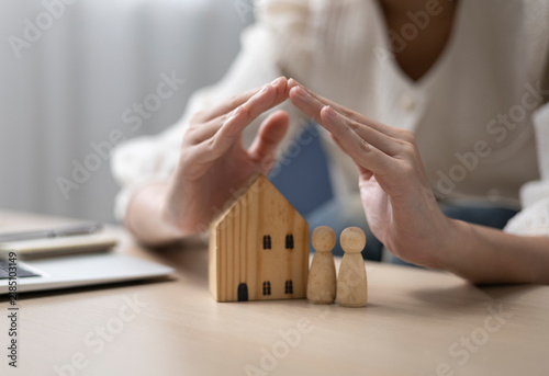 The woman's hands protecting the wooden house and the family portrait symbolize property protection and financial stability. Home insurance, family protection, property security.