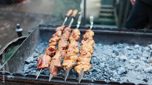 Skewers of marinated meat grilling on charcoal barbecue. Shashlik cooking over hot coals on metal grill. Traditional outdoor barbecue street food preparation concept.