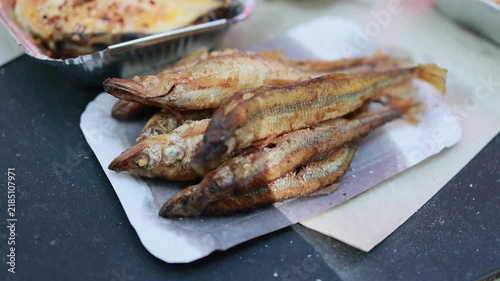 Fried small fish served on paper tray at street food market. Grilled smelt snack on dark table with baked dish in foil container. Traditional street cuisine and outdoor eating concept