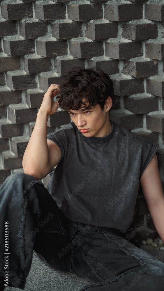 Obraz premium Teenage boy runs a hand through his hair while sitting against a geometric concrete wall and looking down, a vertical urban frame about tiredness and thinking things over alone