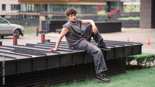Teenage boy leans back on a long black bench on a city street with parked cars and an office building behind him, a horizontal urban lifestyle frame for youth and street brands