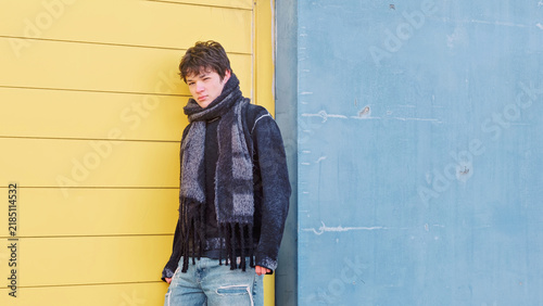 Teenage boy in a denim jacket and a grey scarf leans against a bright yellow and blue painted wall, a colour block street style portrait for youth fashion and social media content