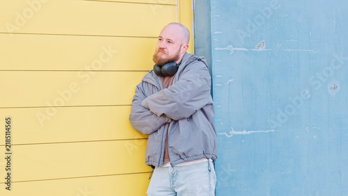Bald bearded man in a denim jacket stands with his arms crossed against a bright yellow and blue painted wall, a confident colour block street style portrait for menswear brands
