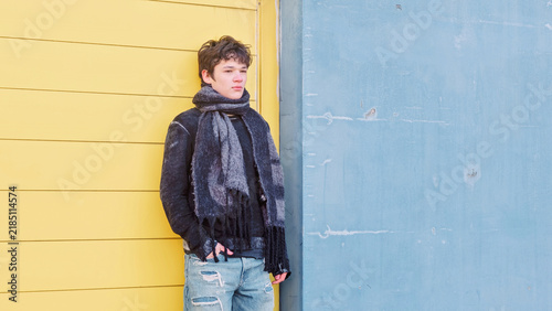 Teenage boy in a scarf and a denim jacket stands against a yellow and blue colour block wall and looks straight into the camera, a bright street style frame for youth clothing brands