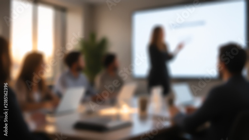 Blurred view of a business presentation: a woman speaking to colleagues by a large screen