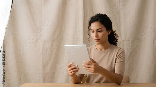 Young woman with curly dark hair sitting at a wooden desk and holding a silver tablet computer against a beige textile background in a bright studio environment.