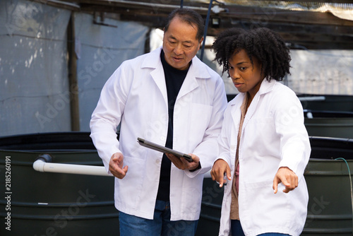 Diverse coworkers in white lab coats examining dark circular tanks and PVC piping using tablet