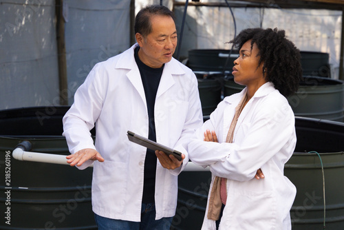 Diverse coworkers in white lab coats inspecting greenhouse tanks with tablet and PVC piping