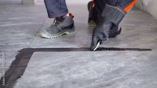 Professional worker in protective gloves applying black sealant to joints on grey ceramic tile floor
