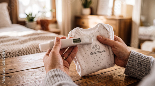 Closeup of female hands holding pregnancy test and baby bodysuit in room