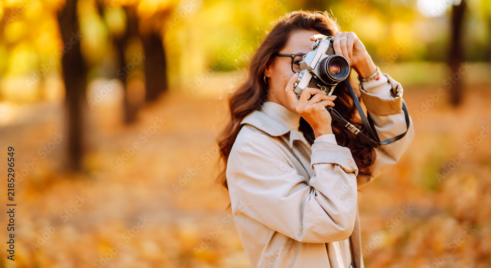 Obraz premium Cheerful woman sits in park surrounded by colorful autumn leaves, holding camera. Woman in coat relaxes in autumn park and takes photos at sunset. Concept of leisure, beauty, and enjoyment.