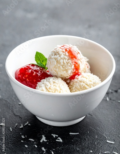 Homemade coconut balls dessert served in a white bowl with raspberry sauce and mint leaves, healthy sugar-free raw vegan Raffaello style candies, minimalist food photography.