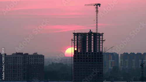 Skyscraper building under construction with tower crane against bright pink sunrise sky time lapse. High rise residential architecture in urban cityscape.