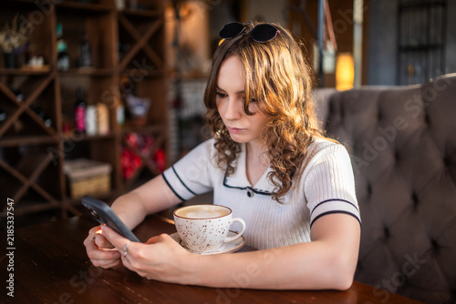 Young woman with curly hair using smartphone with coffee in cafe