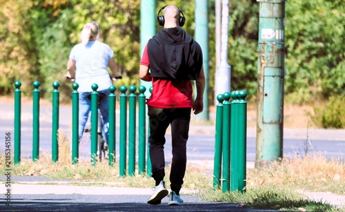 Bald man wearing headphones walking beside green roadside poles