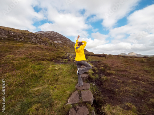 Canvastavla Person in a bright yellow raincoat stands on a stone path, facing a vast, rugged landscape