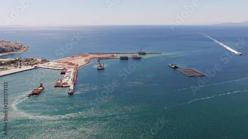 Industrial machinery working on a major port expansion project, including land reclamation and breakwater construction, with vessels maneuvering in the bay and a fast ferry crossing the sea