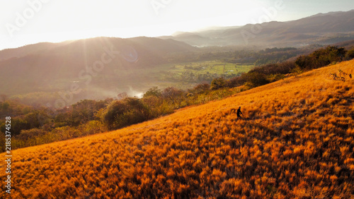 Fotografie A lone hiker walking across a golden grass-covered hillside overlooking a green valley and distant mountain ranges under warm natural sunlight