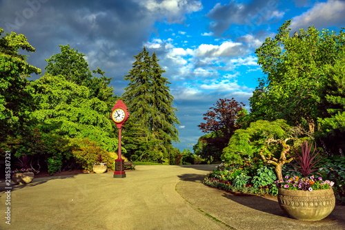 A street clock on a post along the central avenue in Vancouver's Stanley Park, with green trees lining the path and flowerbeds set against a cloudy sky