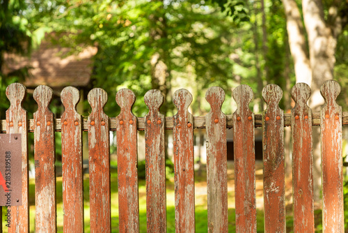 Weathered wooden picket fence with peeling orange-brown paint in front of green trees.