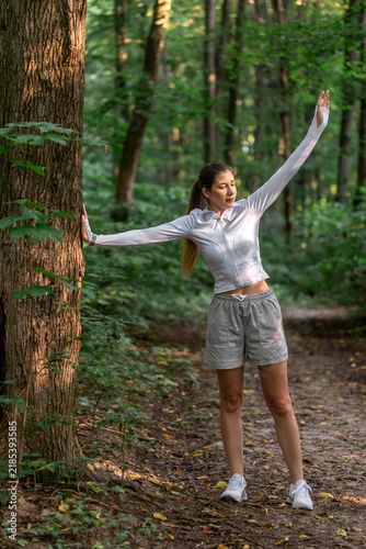 Woman stretching arms beside a tree in forest. Outdoor workout and fitness routine.