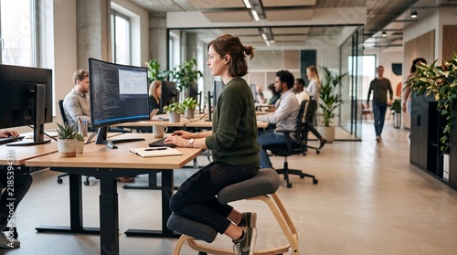 Woman programmer working on computer in modern open plan office with ergonomic kneeling chair