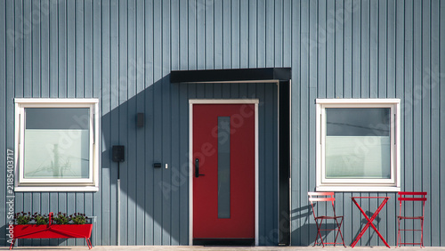Modern house main entrance with bold red front door, dark gray vertical wood cladding, simple black canopy porch, white-framed windows, and red outdoor chairs and table