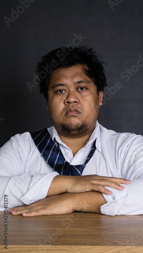 Canvas Print A serious young Asian male professional sits directly facing the camera with folded arms resting on a wooden desk against a pitch black backdrop