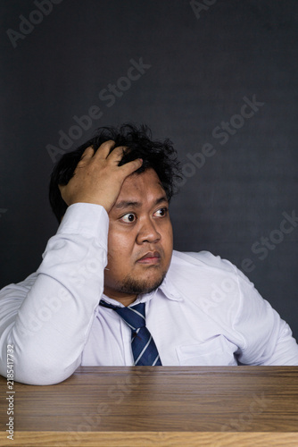 Foto A worried young Asian male professional sits at a clean wooden desk against a solid dark gray wall, clutching the top of the head with one hand while staring anxiously off to the side