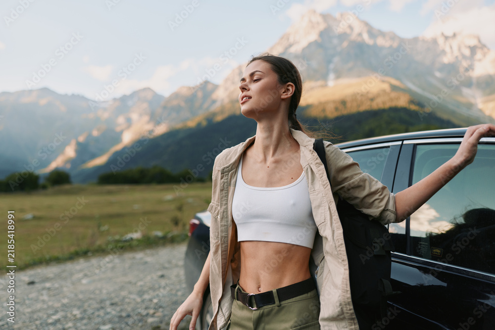 Obraz premium Cheerful woman outdoors, standing by a car with mountains behind, cropped top and light jacket, sunlit breeze, casual lifestyle, carefree moment, vibrant natural colors