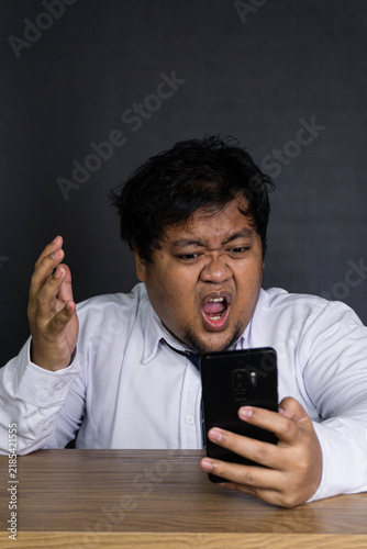 Foto A young Asian male wearing a white business shirt and blue necktie screams in pure rage while aggressively looking at a black smartphone