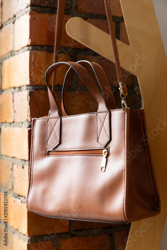 Brown leather tote bag hanging by shoulder strap on wooden coat stand against red-orange brick wall