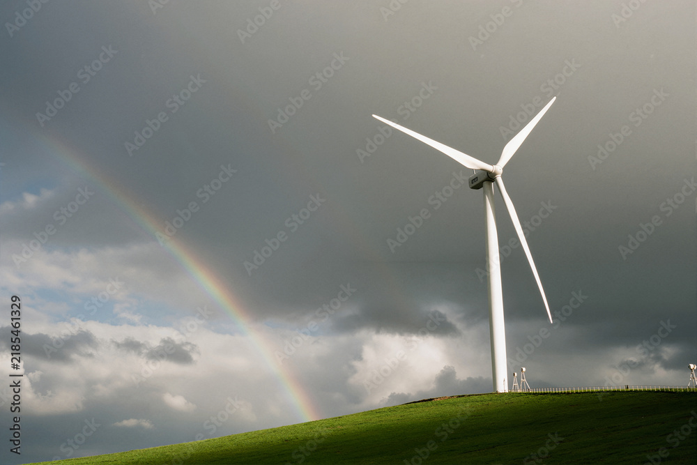 Obraz premium Wind turbine on grassy hill with rainbow against stormy sky
