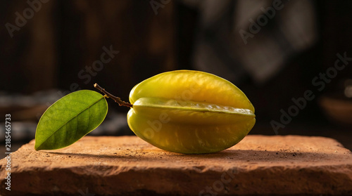 Low-angle macro shot of whole starfruit with green leaf on terracotta slab with warm rim lighting