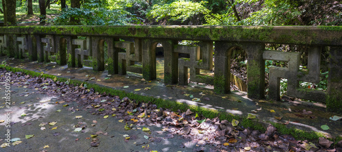 A mossy stone bridge railing decorated with Buddhist manji patterns spans across a forest stream near a Korean temple entrance.