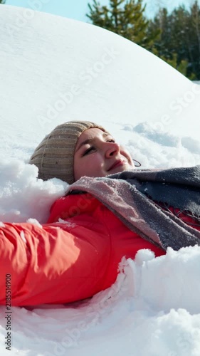 A happy young girl, playfully lying on the white snow, draws an angel with her hands and smiles. People in winter, winter mood. Close-up. Vertical video.