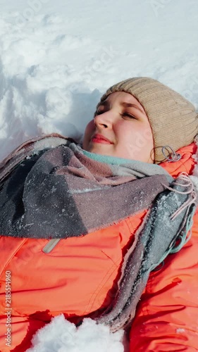 A happy young girl, playfully lying on the white snow, draws an angel with her hands and smiles. People in winter, winter mood. Close-up. Vertical video.