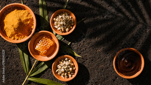 Flat lay of herbal ingredients for tropical health, clay bowl, containing golden turmeric powder, dried jasmine flowers, green pandan leaves, raw honey with natural honeycomb, and coconut oil.