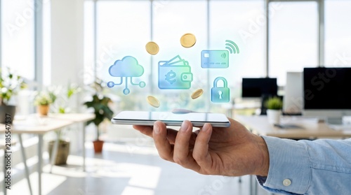 A hand holds a smartphone with digital icons representing cloud computing, digital wallet, credit card, security lock, and currency in a bright office background.