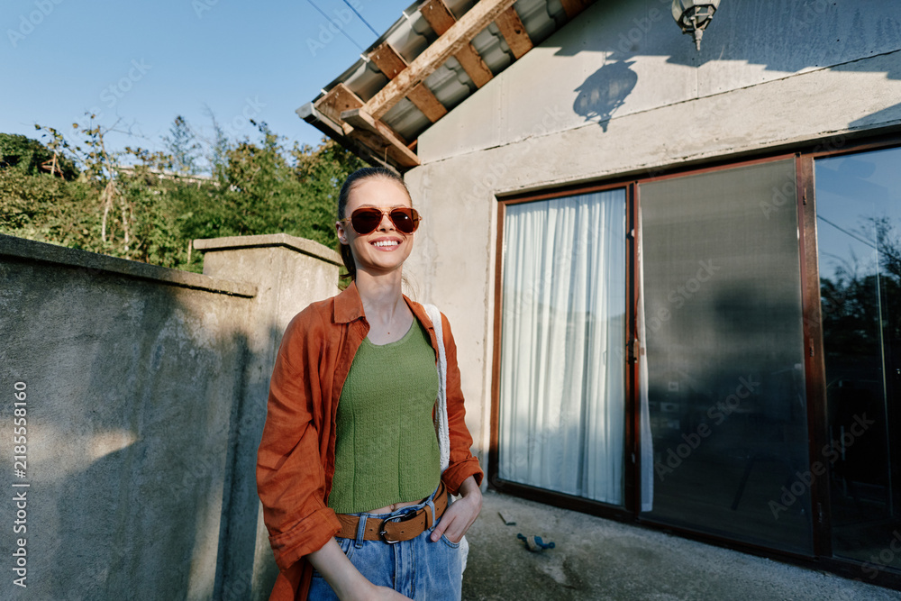 Obraz premium Smiling young woman in sunglasses and orange shirt stands outdoors near a rustic house wall. Casual summer style, travel lifestyle and relaxed vacation mood concept.