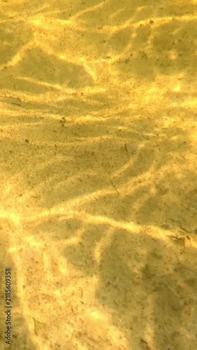 Underwater shot of textured golden sand seabed with shifting light and shadow patterns, sunlight ripples move steadily across the shallow seafloor as camera slowly pans over.