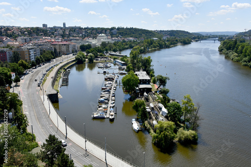 Bild auf Leinwand A wide high-angle view of the Vltava River in Prague featuring a curved embankment, a small island, and docked boats along a marina under a clear sky
