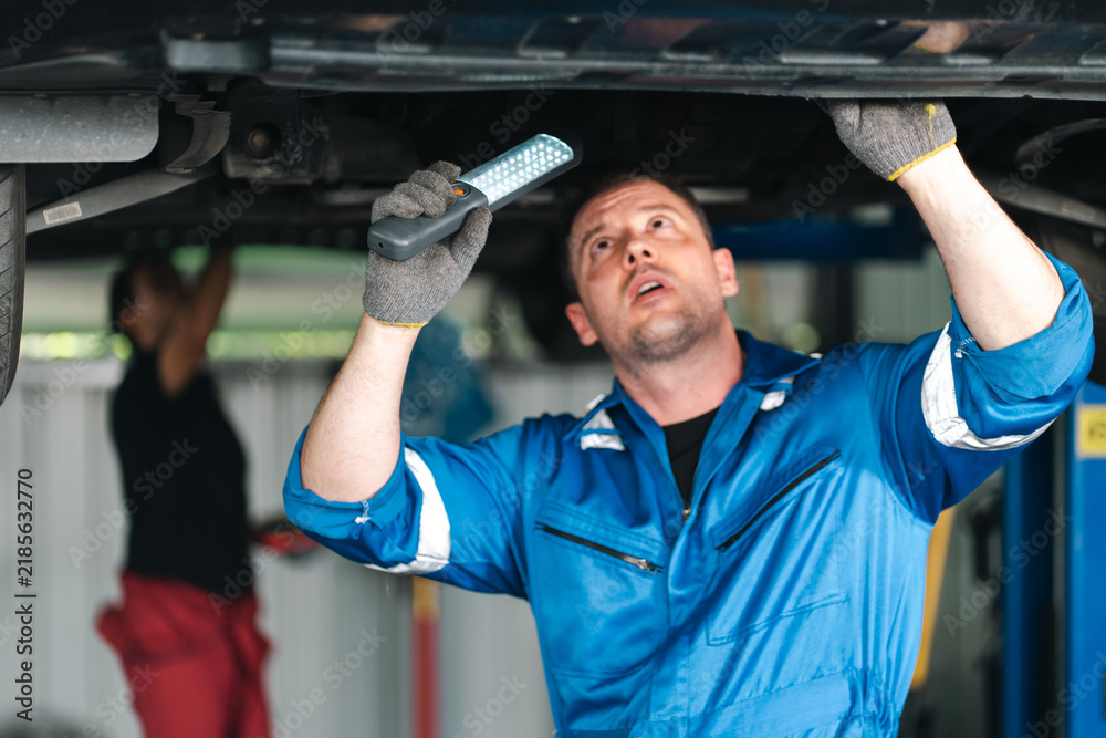 Obraz premium Ev car battery cells service Automotive mechanic inspecting vehicle underside with flashlight in repair garage, focused male technician wearing blue workwear and protective gloves