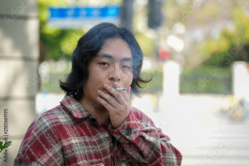 A person holds a cigarette near the mouth while standing outdoors on a city sidewalk.