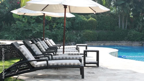 Rows of lounge chairs face a calm swimming pool. Large umbrellas shade the seating area. Lush greenery surrounds the poolside. Sunlight glints off the clear water