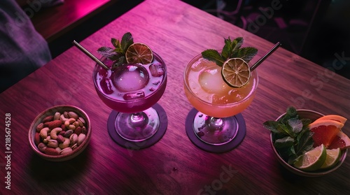Top view of two colorful cocktails served with nuts and fresh citrus garnishes on a wooden table under vibrant pink neon lighting