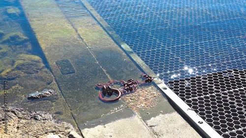 rusty metal chain and ring, partially submerged in shallow water at the edge of a concrete ramp or dock. river Orb. Valras, France