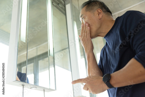 Senior Asian man covering mouth while pointing at mirror cabinet by window in washroom, copy space